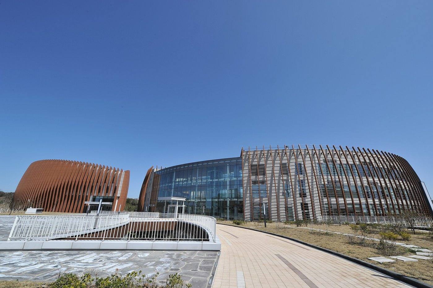 Branksome Hall Asia in Jeju Global Education City: a curved glass-fronted building and a long façade wrapped in vertical rust-red fins, with a second fin-clad building on the left and a paved path curving toward the entrance under a deep blue sky, March 2013.
