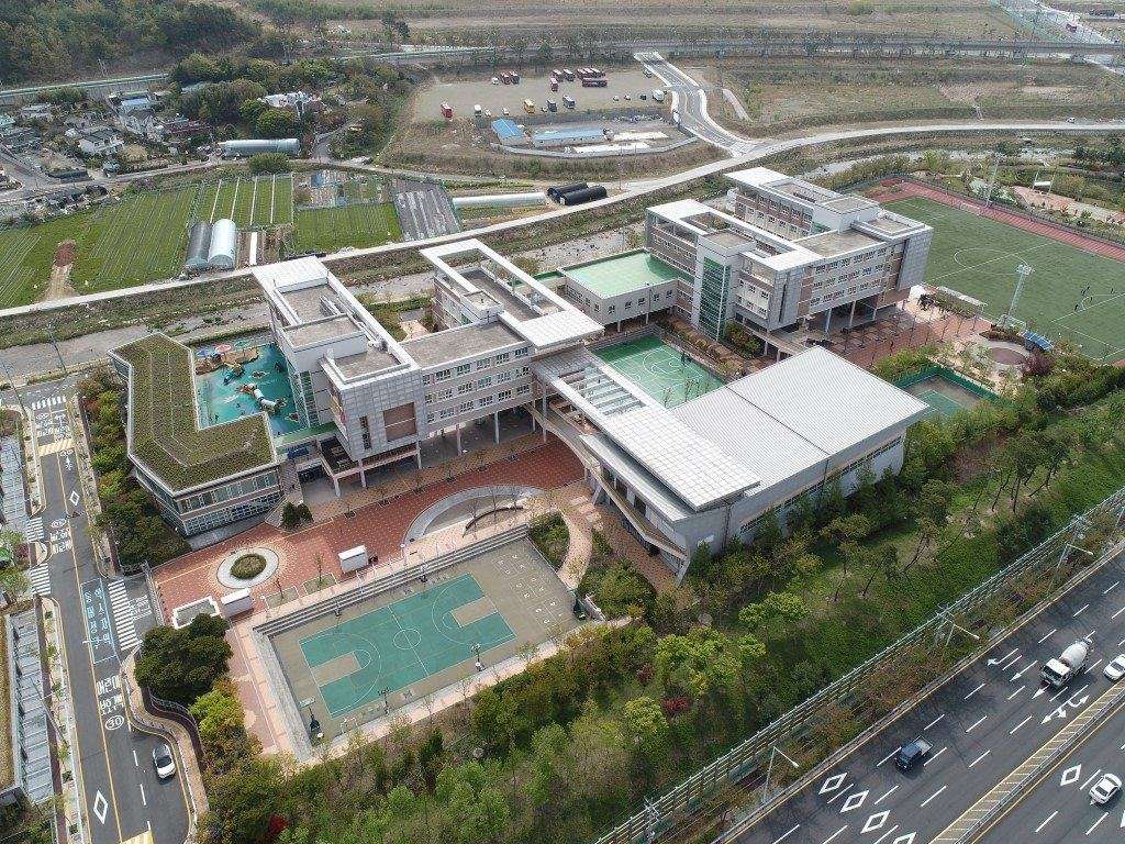 An aerial view of the International School of Busan: a cluster of white and grey school buildings with a green roof garden, a covered courtyard, outdoor basketball courts and a full-size football pitch beside a highway, with farm fields and hills behind, 2019.