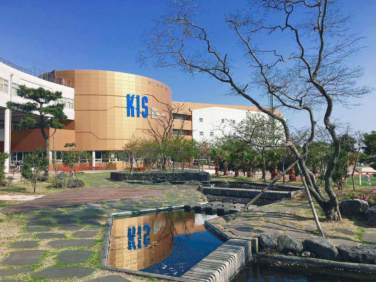 The campus of Korea International School Jeju in Jeju Global Education City: a curved orange-gold building with large blue "KIS" letters, white wings on either side, a lawn with stepping stones and a reflecting pool in front, and bare trees under a clear blue sky, April 2017.