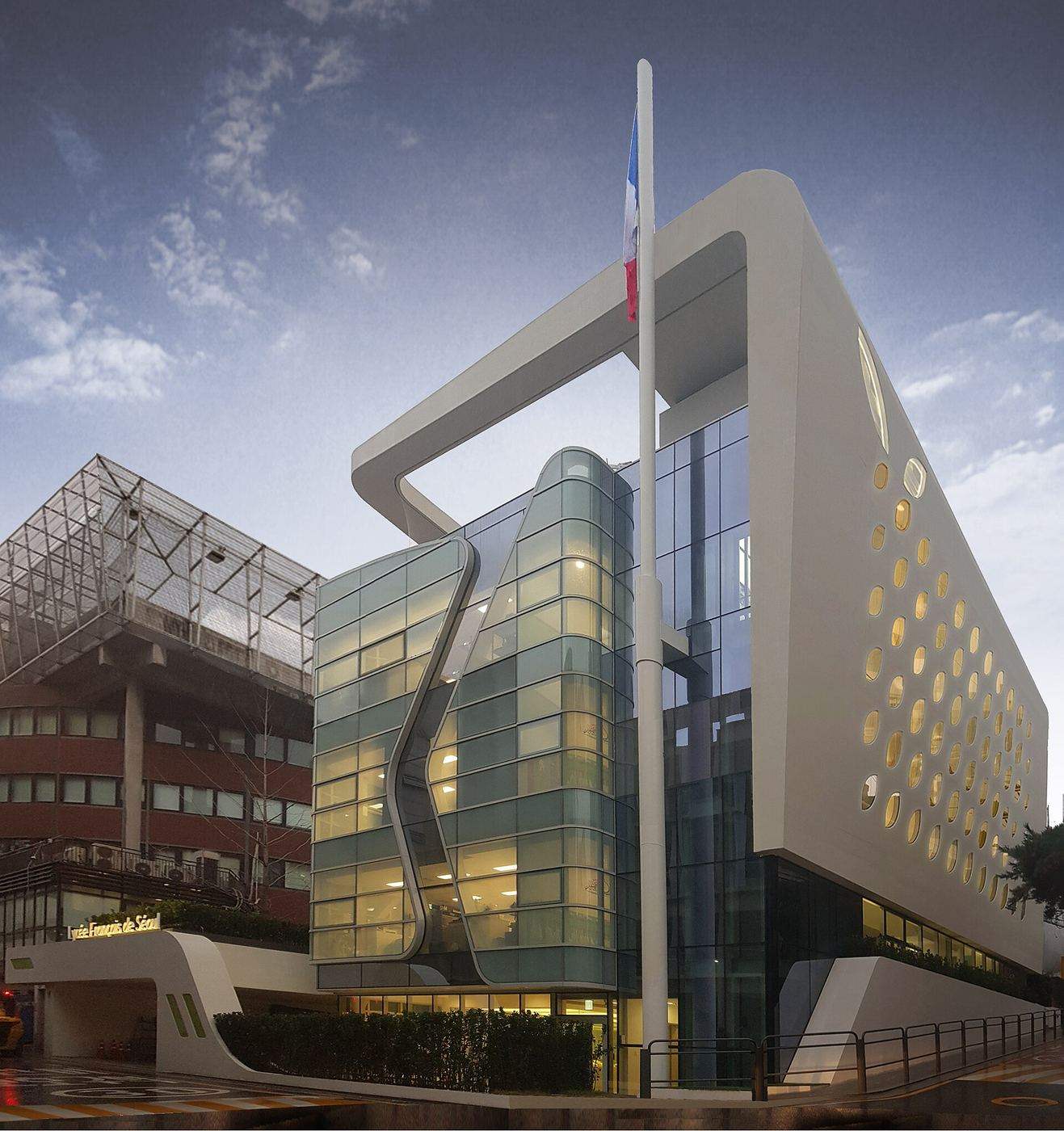 The Lycée Français de Séoul in Seocho-gu, Seoul: a modern white building with a large looped frame across its top, porthole-shaped windows on one side and a curved glass façade, a French flag on a tall pole in front, and the school's name on a low canopy at the entrance, June 2019.