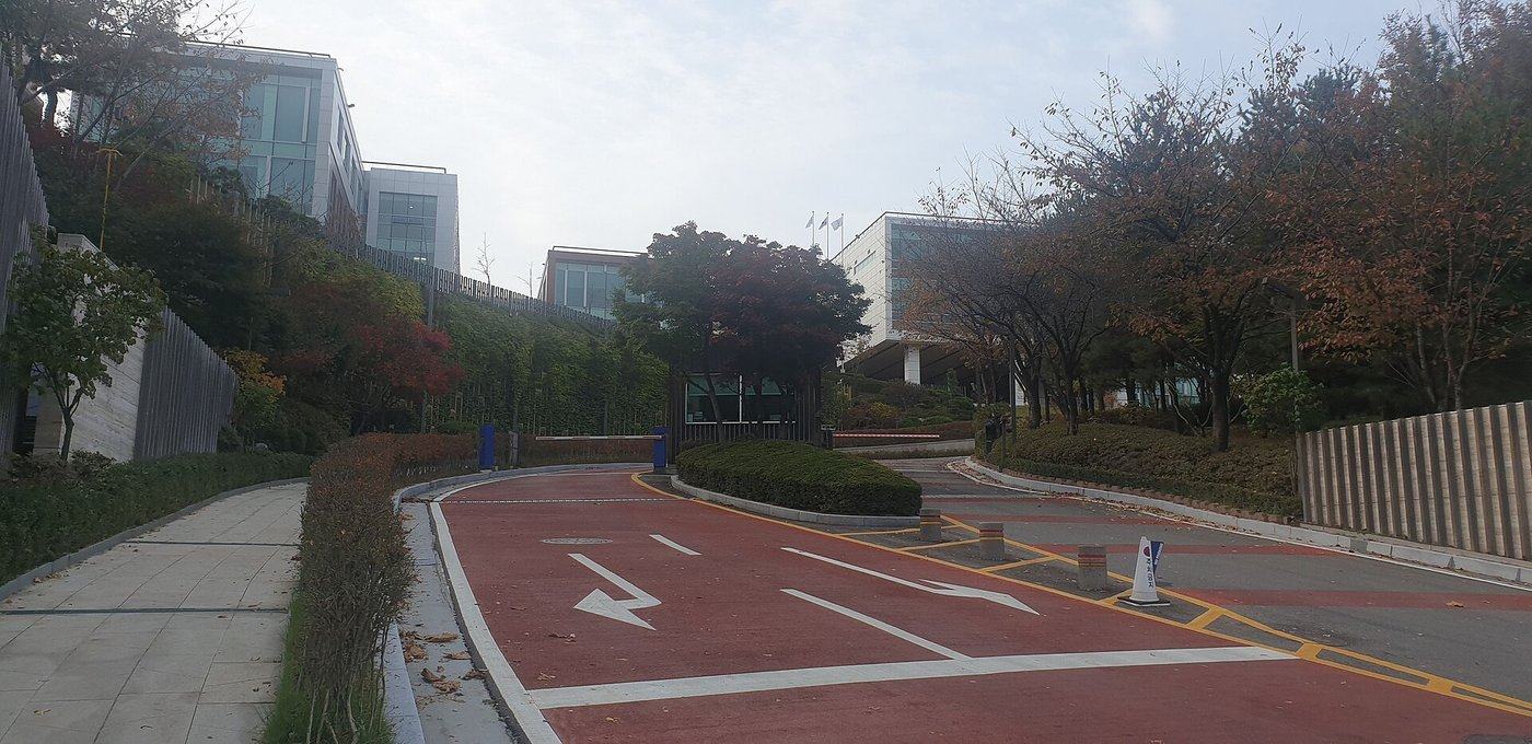The entrance road of Yongsan International School of Seoul: a red-surfaced driveway with white arrows rising to a security barrier, glass-and-white school buildings on the hillside above, and autumn trees in red and brown on both sides, November 2023.