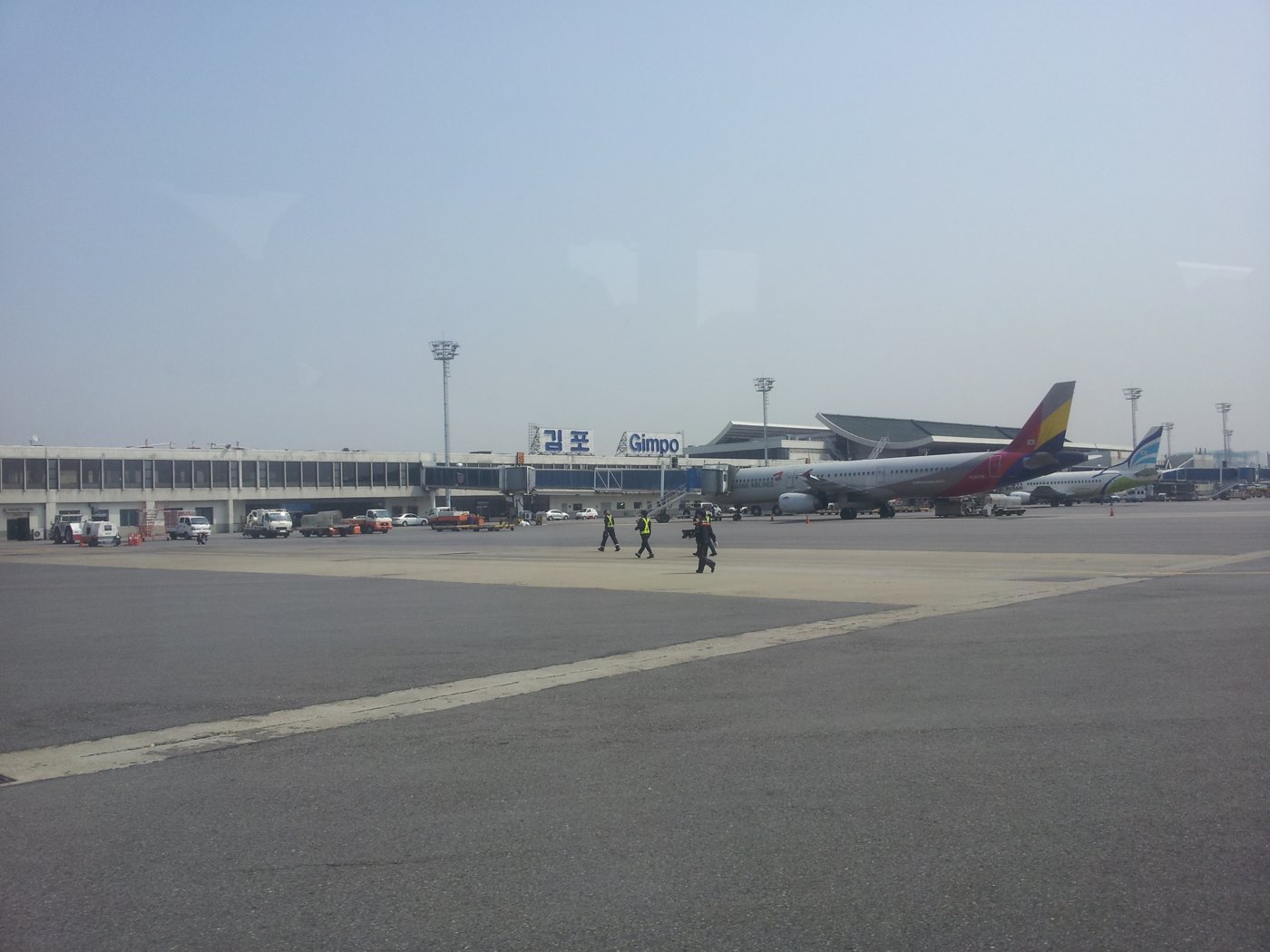 The apron at Gimpo International Airport seen from a taxiing aircraft: a long low terminal with the signs 김포 and Gimpo on the roof, an Asiana Airlines Airbus parked at a gate with its multicoloured tail, another airliner behind it, ground vehicles and ground staff in high-visibility vests walking across the tarmac under a hazy sky.