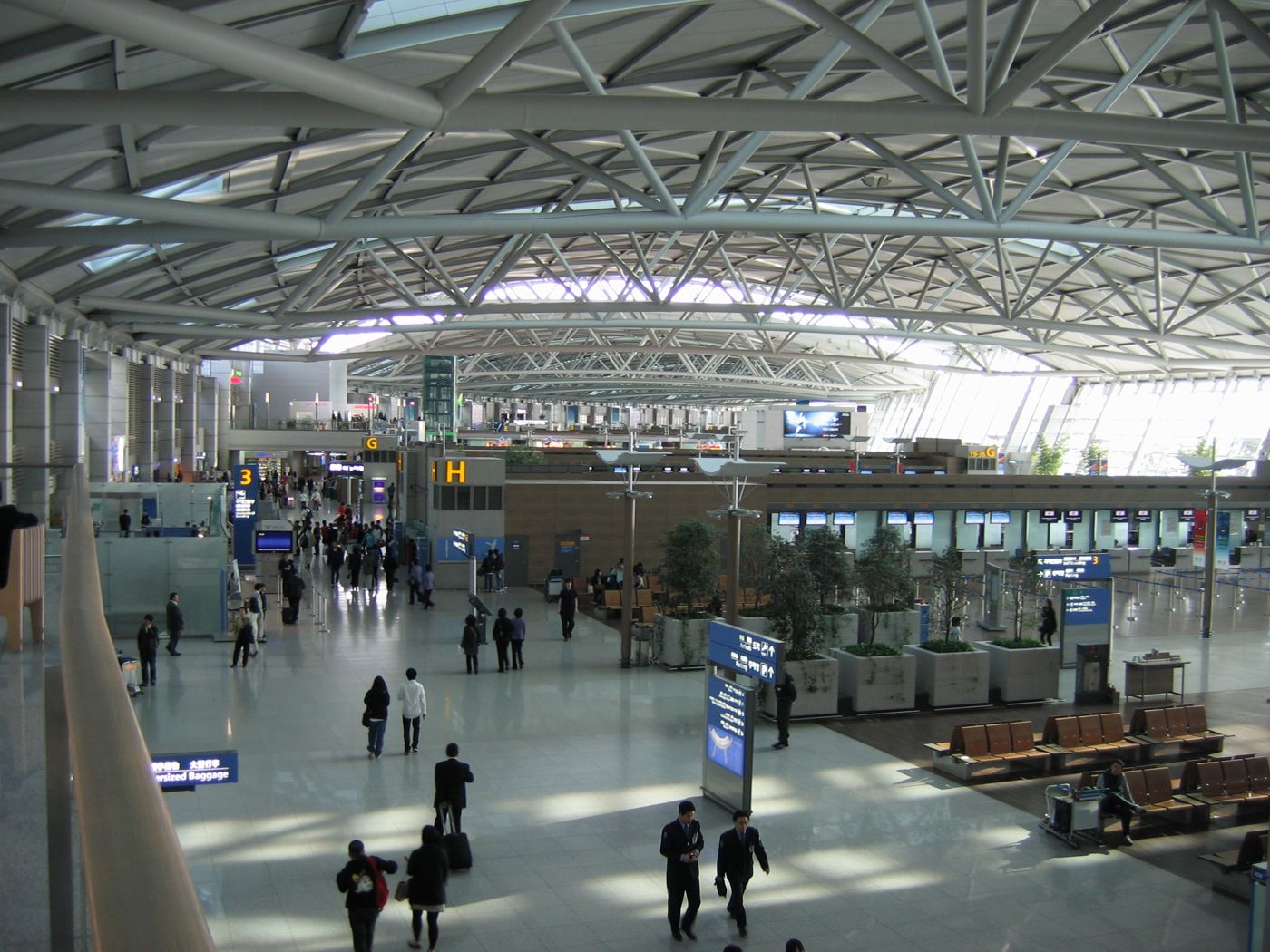 The departures hall of Incheon International Airport Terminal 1 seen from an upper walkway: a long steel truss roof, check-in island H on the right with blue signs and rows of brown seats, potted trees, and passengers with suitcases walking across a polished floor.