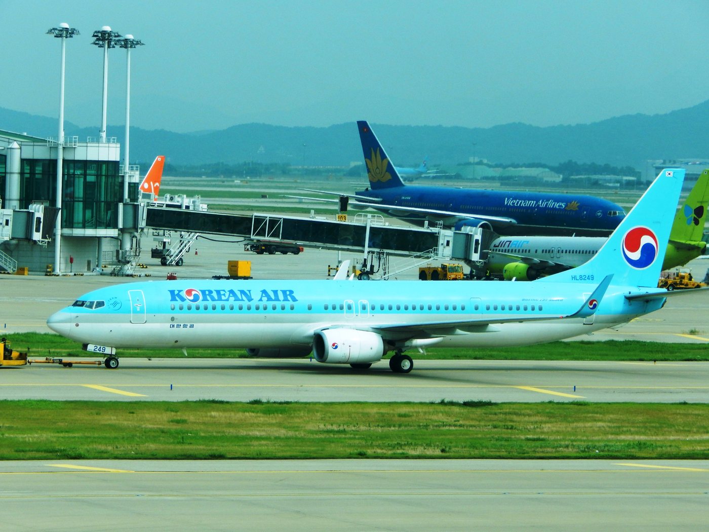 A Korean Air Boeing 737-900 in the airline's light-blue livery with the red and blue taeguk tail, registration HL8249, being towed along a taxiway at Incheon Airport; behind it a Vietnam Airlines widebody and a Jin Air 737 sit at the midfield concourse.