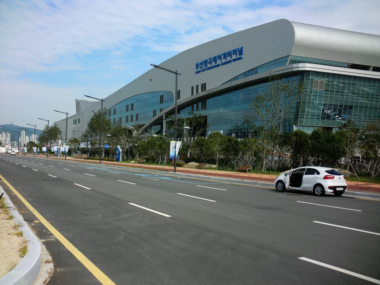 Busan Port International Passenger Terminal seen from the road in front: a long, low glass building with a curved silver roof and the Korean name 부산항국제여객터미널 in blue letters, young trees and a wide empty road in the foreground, October 2015.