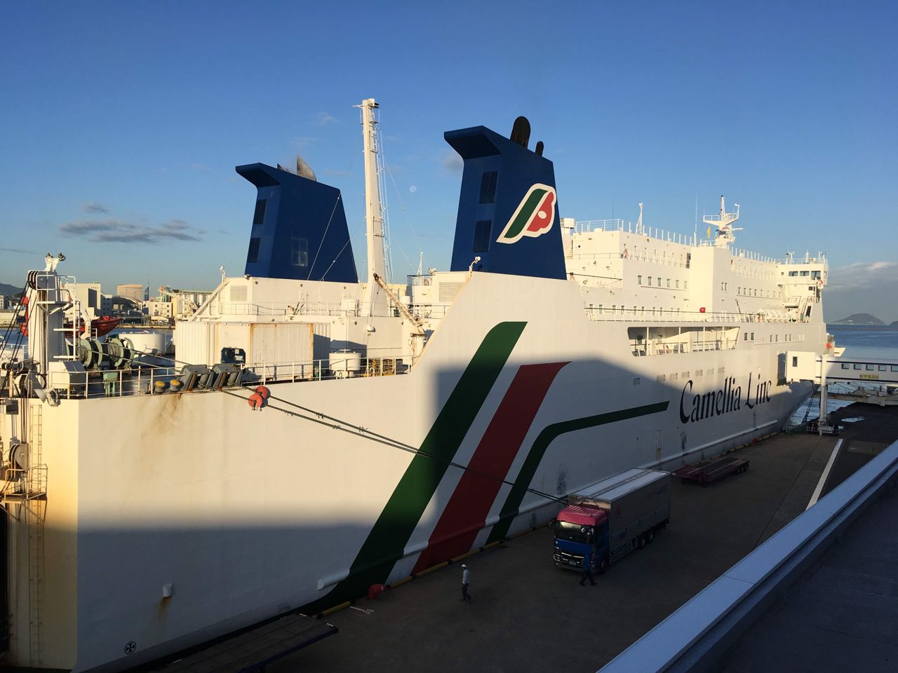 The New Camellia, white with green and red stripes and "Camellia Line" on the side, berthed at Hakata Port in morning sunlight, seen from a walkway above the quay, with a truck on the dock and the ship's blue funnel with a red-and-green logo, September 2018.