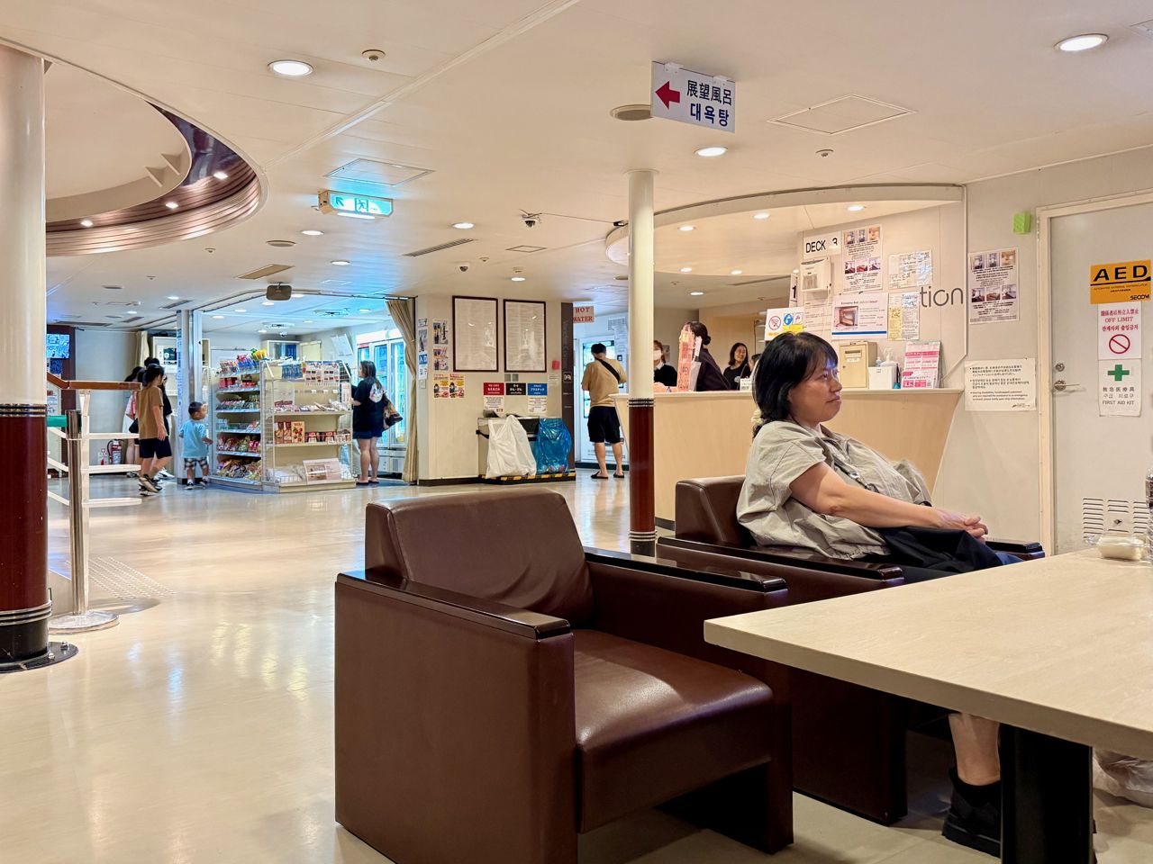 The lobby of the New Camellia: brown leather armchairs and a table in front, a small shop with snacks and drinks behind, an information desk to the right, and a hanging sign with an arrow reading 展望風呂 and 대욕탕 for the bath, July 2025.