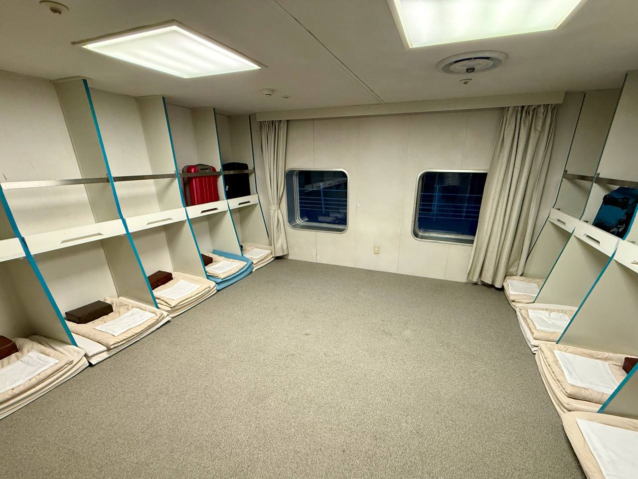 A shared second-class room on the New Camellia: grey carpet with thin white mats and brown pillows laid along both sides, open shelving above the sleeping spaces with suitcases on it, two portholes with curtains at the far end, July 2025.