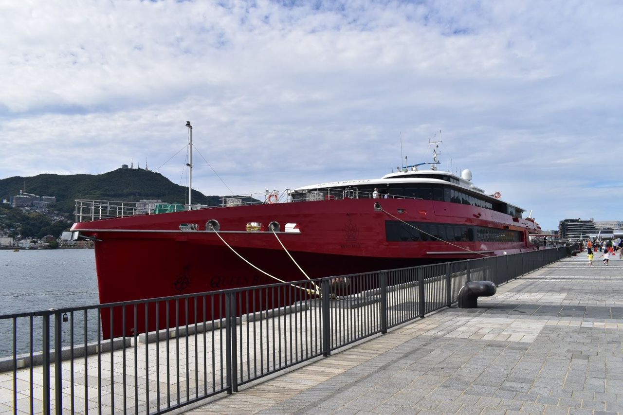 The red trimaran Queen Beetle moored alongside a quayside promenade with railings and a bollard, hills with buildings behind and people walking at the far end, September 2022.