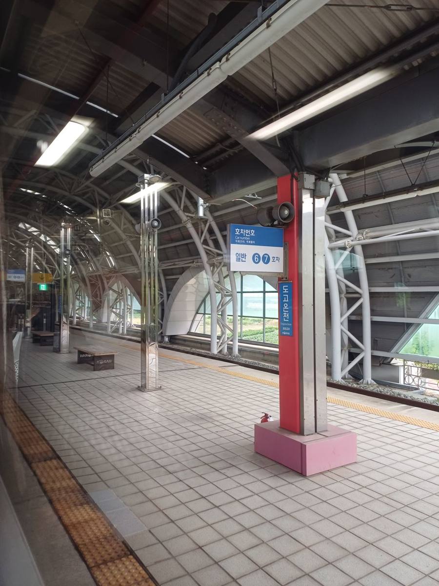 A covered railway platform with arched steel ribs, tiled floor and a red pillar carrying a blue sign reading 도고온천 and a car-number sign, with green fields visible through the side windows.