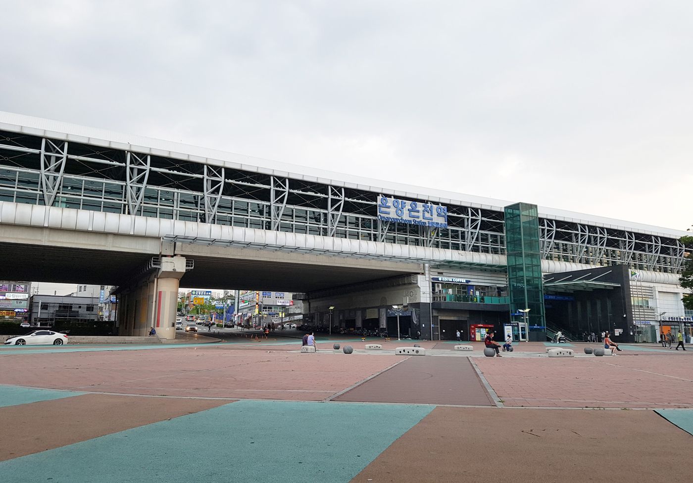 A long elevated station building with a glass and steel roof over the tracks and the blue sign 온양온천역, seen from a wide paved plaza with a few people sitting on stone benches under a cloudy evening sky.