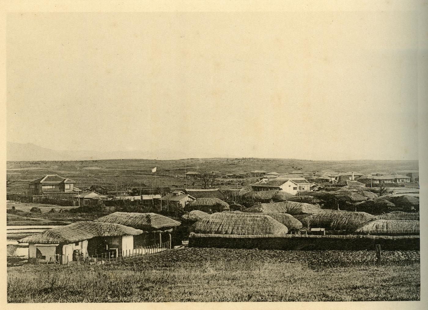 A wide black-and-white view of a flat village of thatched-roof houses and a few tiled, two-storey buildings, with a flag on a pole and low hills on the horizon: Onyang hot spring village in 1912.