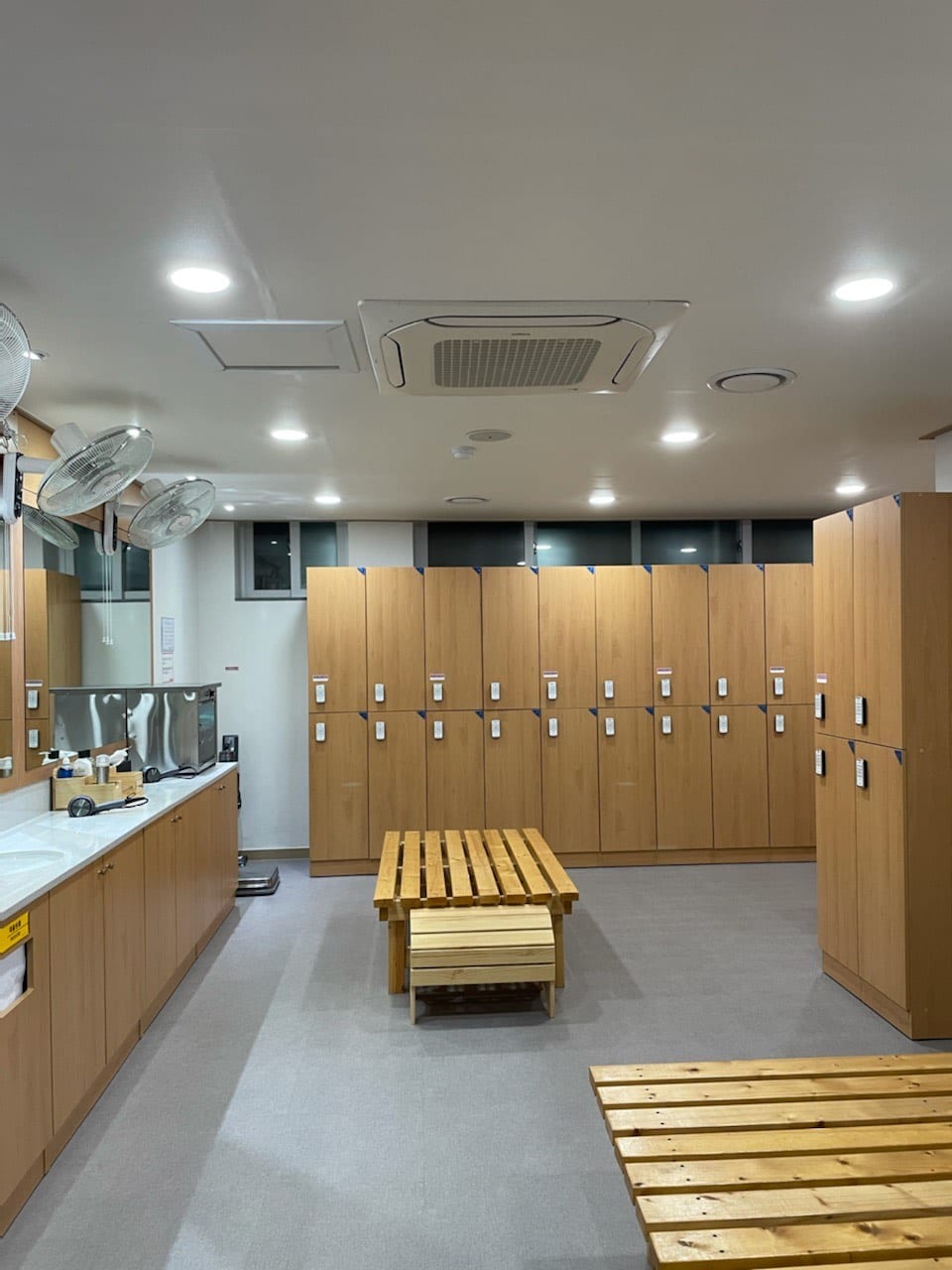 A clean bathhouse changing room with rows of wood-panelled lockers, a long counter of sinks with wall fans above it, and slatted wooden benches in the middle of the floor.