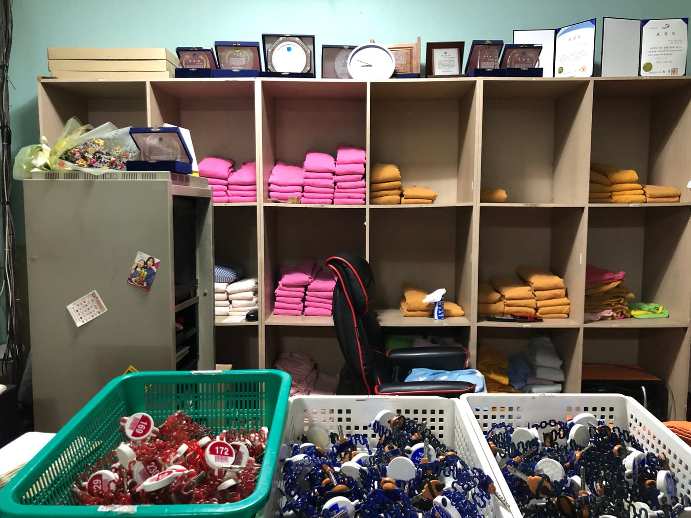 A bath-and-sauna front-counter area: shelves stacked with folded pink and yellow sauna clothes and white towels, and in front, baskets full of numbered locker keys on coloured wristbands.