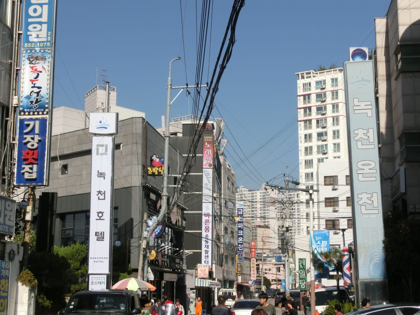 A street in the Dongnae hot-spring district of Busan, November 2018: on the right a tall vertical sign reading 녹천온천 and NOCCHEON HOT SPRING under a wave logo, on the left a matching sign for 녹천호텔 (Nokcheon Hotel), with shop signs and power lines in between.
