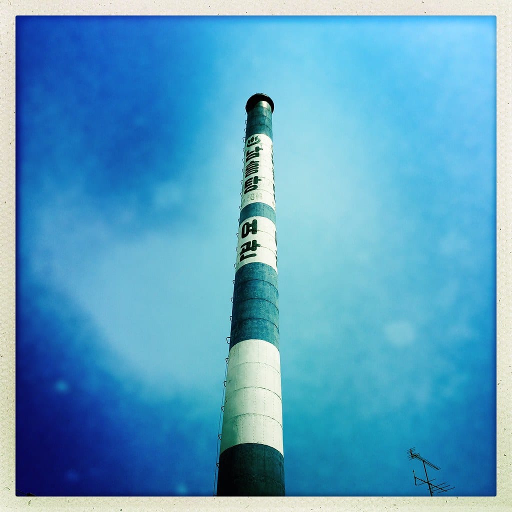 A tall green-and-white bathhouse chimney against a blue sky, painted vertically with the hot-spring symbol ♨, a name ending in 탕 ("bath"), and below it 여관 ("inn").
