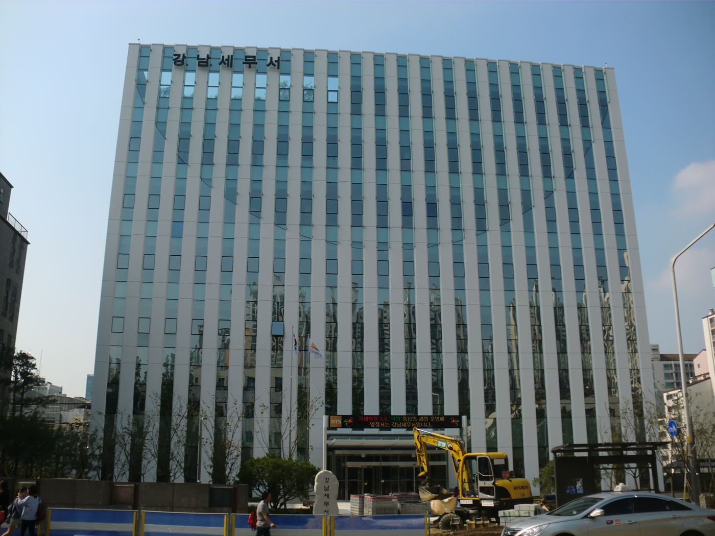 The Gangnam Tax Office in Seoul: a white office building with tall vertical glass strips, the name 강남세무서 in black letters at the top, an LED message board over the entrance, a Korean flag and an excavator on the forecourt.