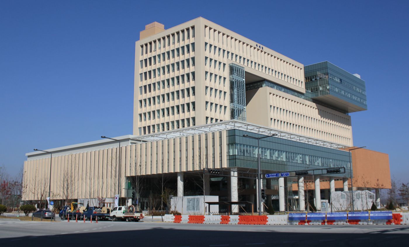 The National Tax Service headquarters in Sejong, South Korea: a cream-coloured office block with a glass bridge and a cantilevered glass wing on the right, the name 국세청 in small letters near the top, and orange-and-white road barriers along the street in front.