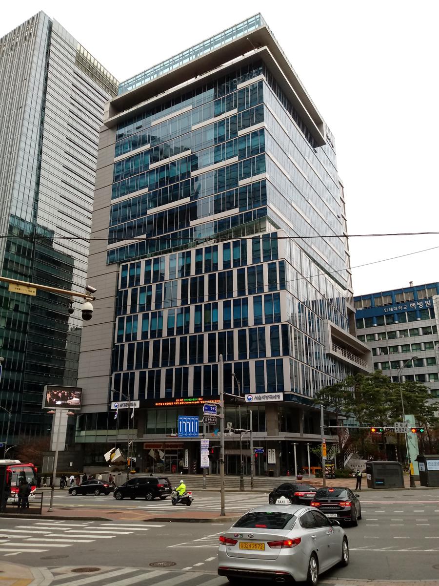 A glass office tower at a Seoul crossroads, with blue signboards over the entrance reading 서울지방국세청 (Seoul Regional Tax Office) and 남대문세무서 (Namdaemun Tax Office), a silver taxi and a motorcycle courier crossing in front.