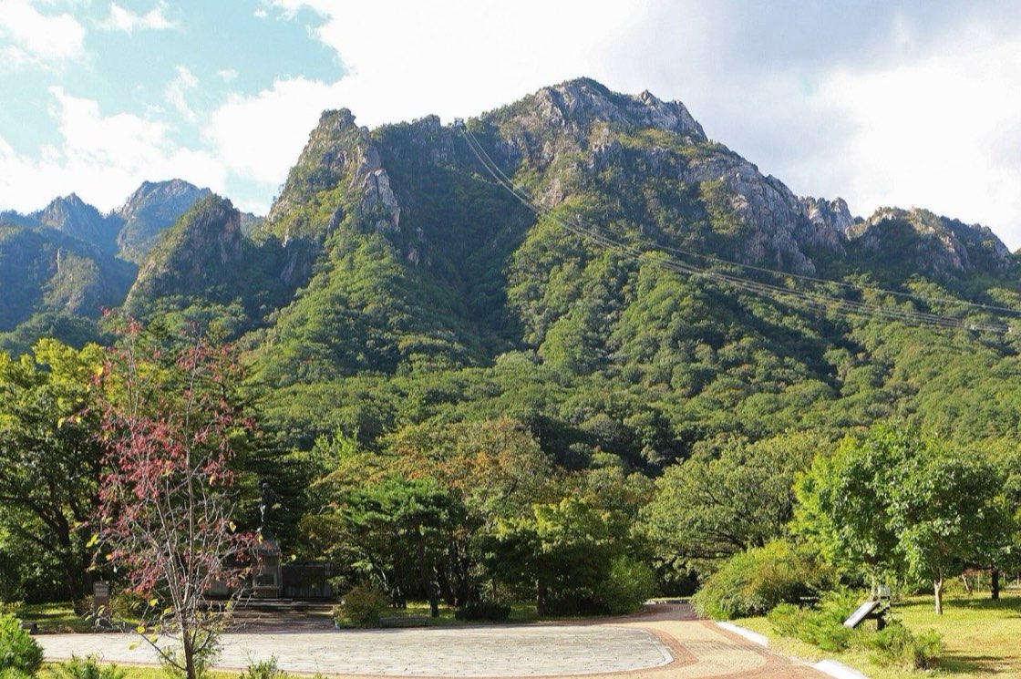 The cable car line running up a steep wooded ridge towards rocky granite peaks above Sogongwon in Seoraksan National Park, with a paved plaza, a young tree with red leaves and green parkland in the foreground, October 2022.