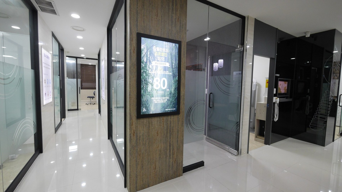 A corridor in a dental hospital in South Korea: glossy white floor tiles, frosted-glass treatment rooms on both sides, a wood-panelled pillar with a backlit poster in Korean, and a treatment chair visible through an open door at the far end.