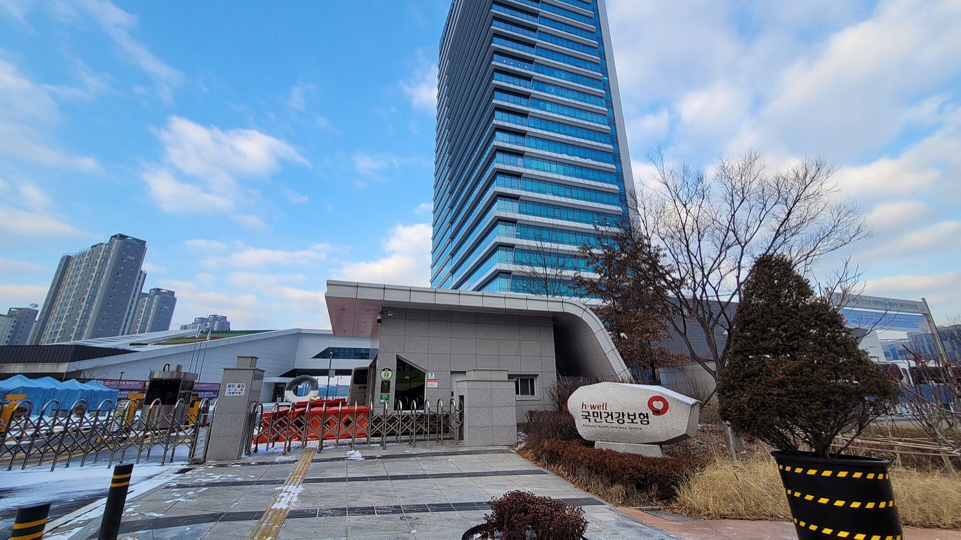 The National Health Insurance Service headquarters in Wonju, South Korea: a tall blue glass tower behind a curved low entrance building, with a white stone marker in front reading 국민건강보험 in red and black, under a blue winter sky with snow on the paving.