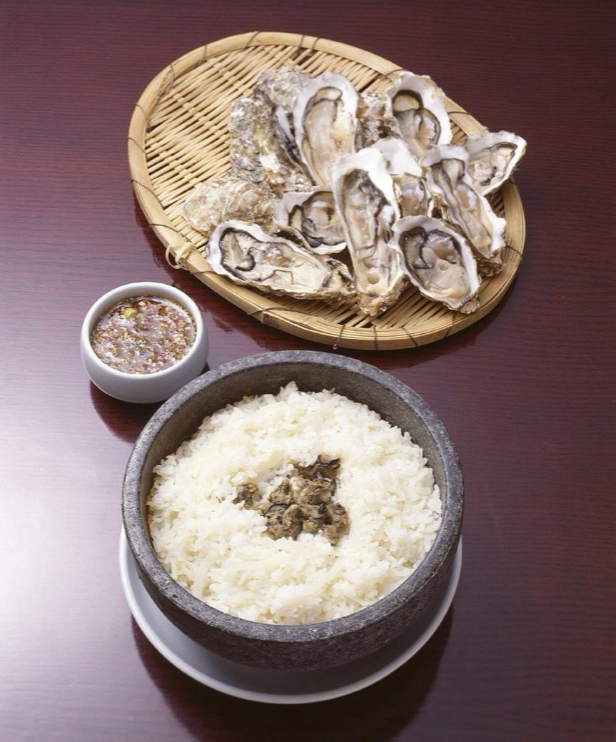 Steamed rice in a dark stone pot with a small heap of cooked oysters in the centre, a bowl of soy and sesame sauce and a bamboo tray of oysters on the half shell behind it.