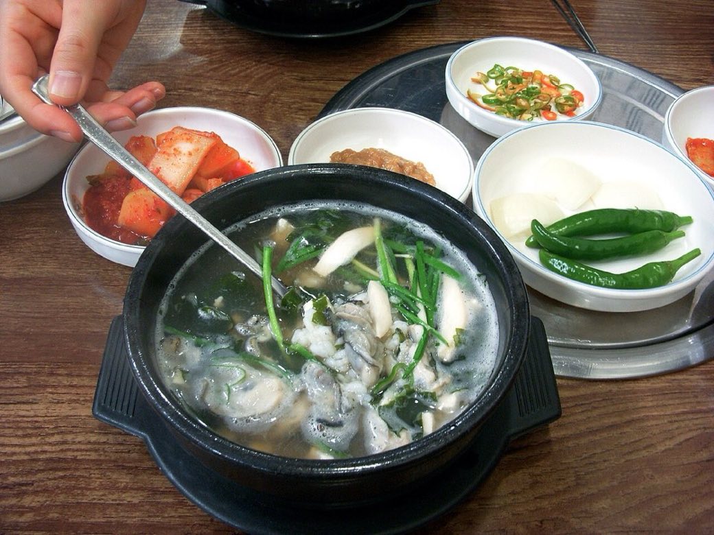 A black stone-pot bowl of oyster rice soup with spring onions and seaweed in clear broth, a spoon lifting it, surrounded by side dishes of kimchi, green chillies, raw garlic and sliced radish on a restaurant table.