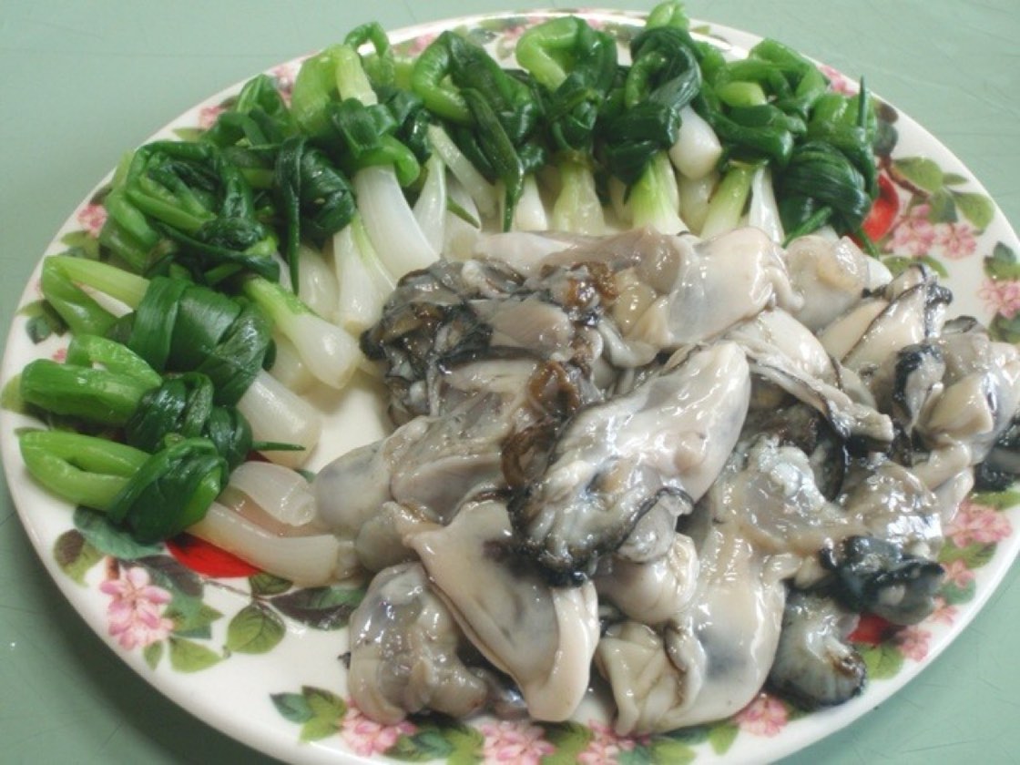 A plate of raw shucked oysters, grey and glistening, beside bundles of blanched spring onion tied in knots on a floral plate, a Korean dish of raw oysters with jjokpa-ganghoe.