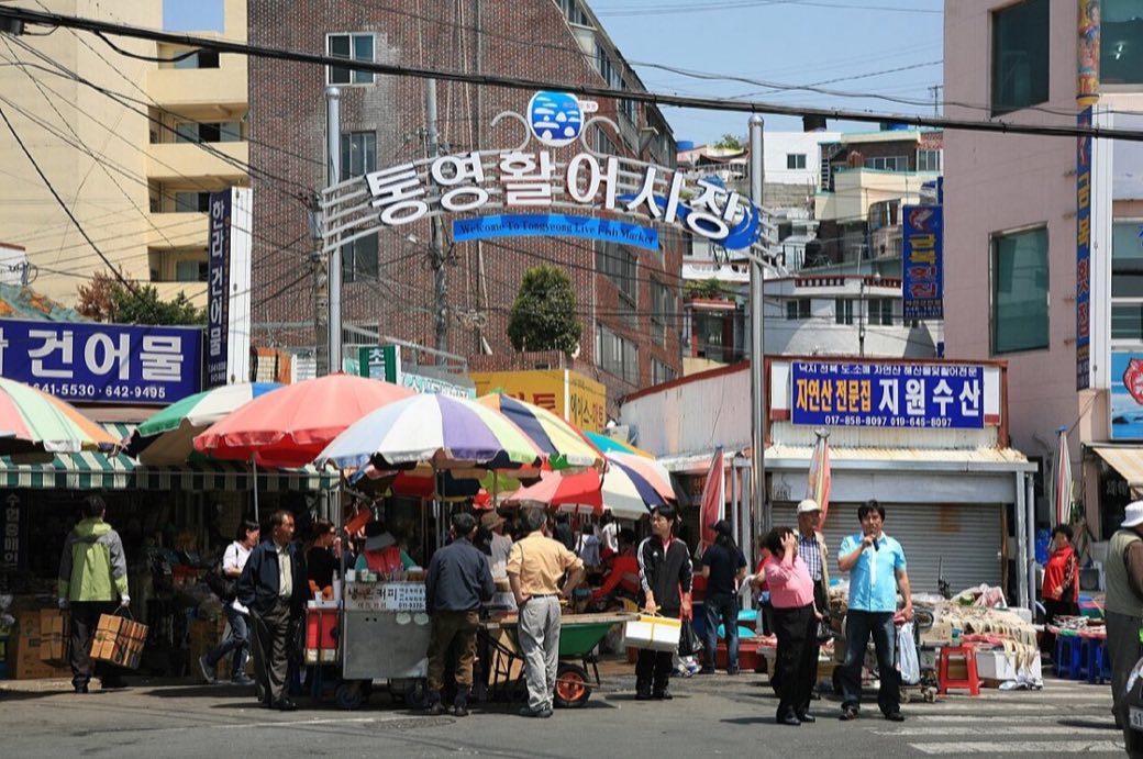 The gateway of Tongyeong's live fish market, a metal arch with the sign 통영활어시장, over a street of beach umbrellas, stalls and shoppers in front of low concrete buildings, May 2009.