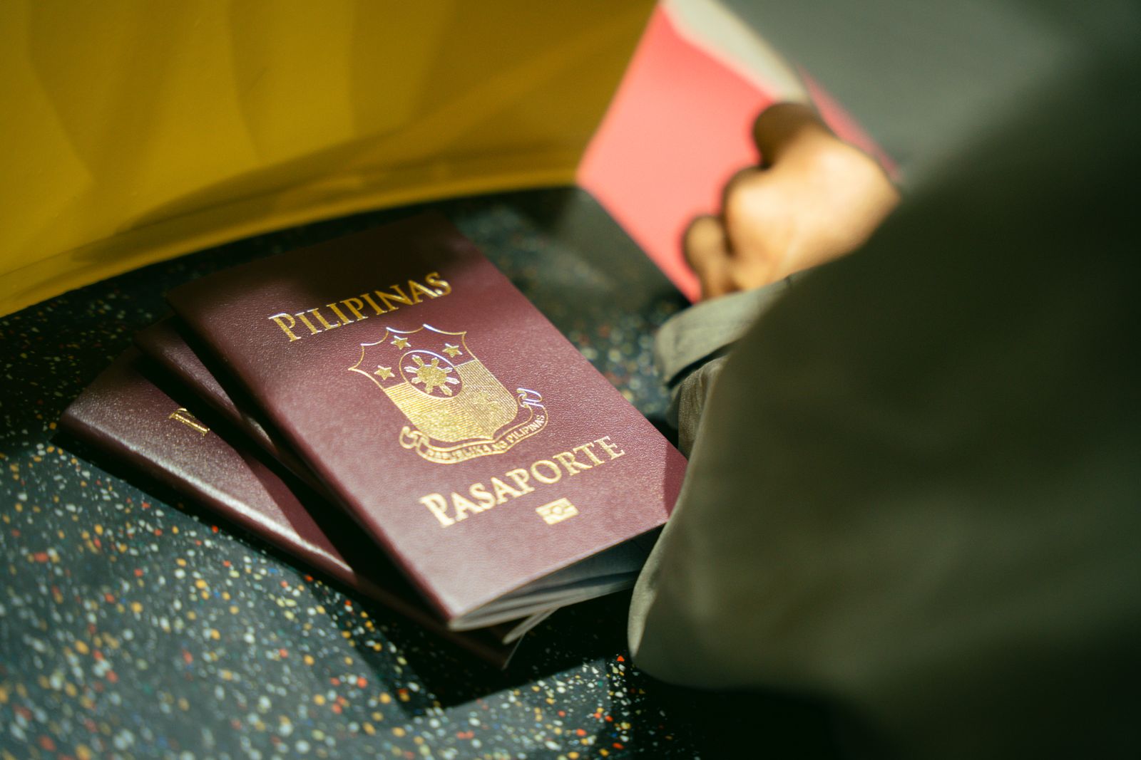 Three burgundy Philippine passports, the top one embossed PILIPINAS and PASAPORTE in gold, lying on a speckled airport floor beside a traveller's knee.