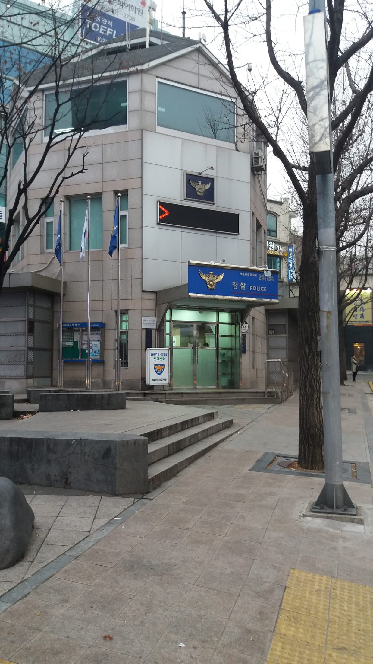 A neighbourhood police box on a Seoul street: a stone-faced building with a blue sign reading 경찰 POLICE and the police emblem over a glass door, flagpoles out front, and a white standing sign beside the door that reads 실종아동·유실물 신고센터 (missing children and lost property reporting centre).