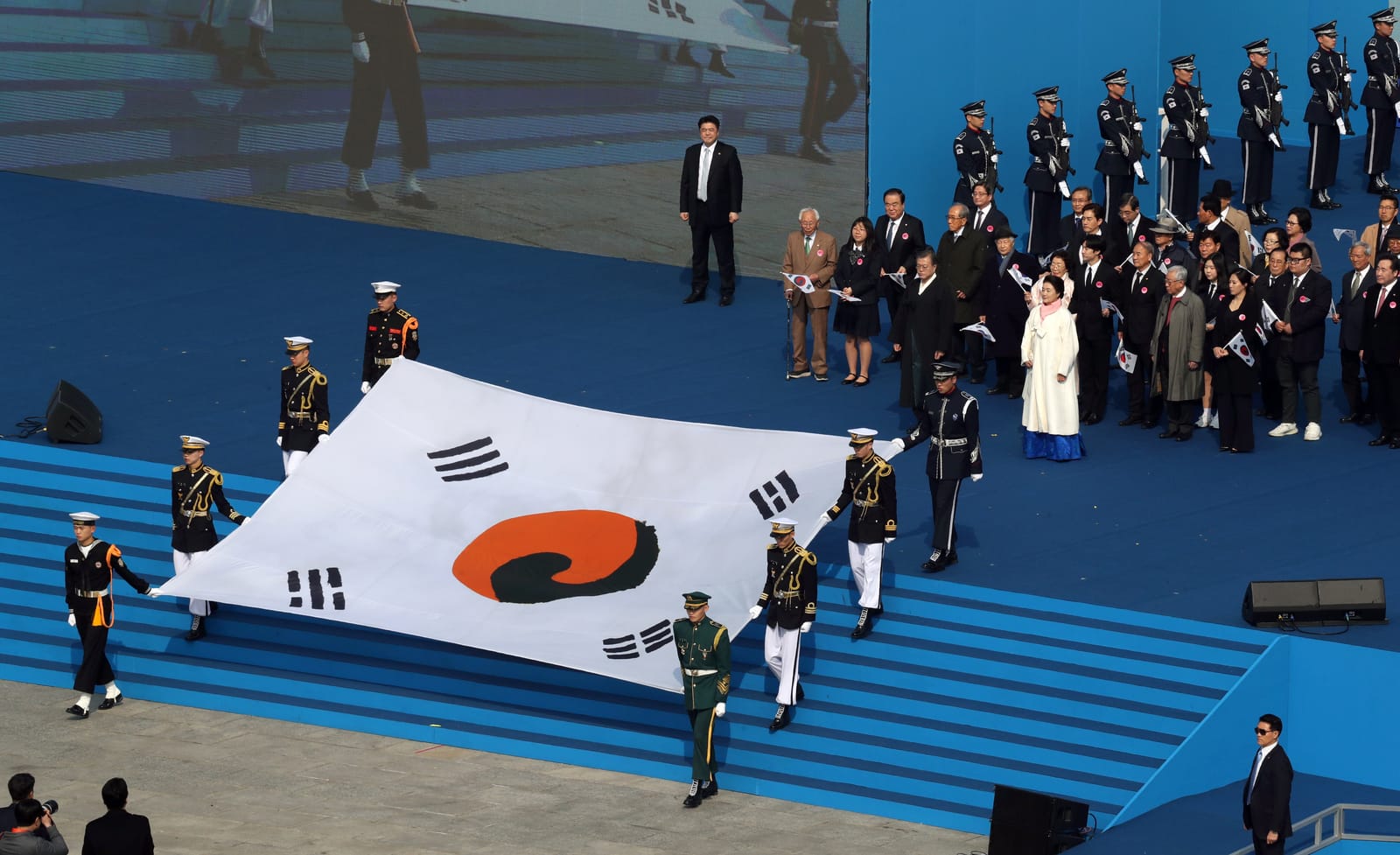 A large Korean national flag, the Taegukgi, carried by a ceremonial honour guard in white-and-navy uniforms down blue steps at Gwanghwamun Square, with officials and a woman in a white hanbok standing behind them holding small flags.
