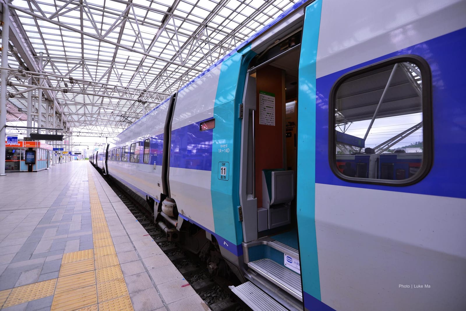 A blue-and-white KTX high-speed train standing at a platform in Seoul Station under a white steel-frame canopy, with the door of car 11 open, showing the step and a folded seat inside, and the platform empty in the morning light.