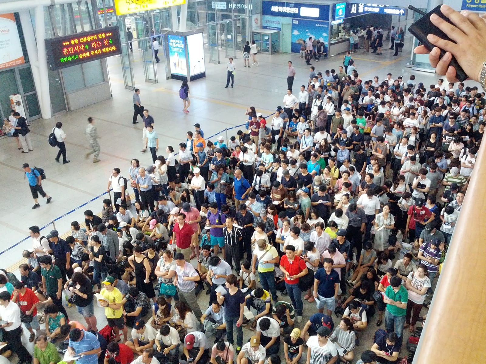 A packed crowd of travellers standing and sitting on the floor of the Seoul Station concourse, filling the hall from wall to wall, while a few people walk past on the left under an orange LED departure sign in Korean.
