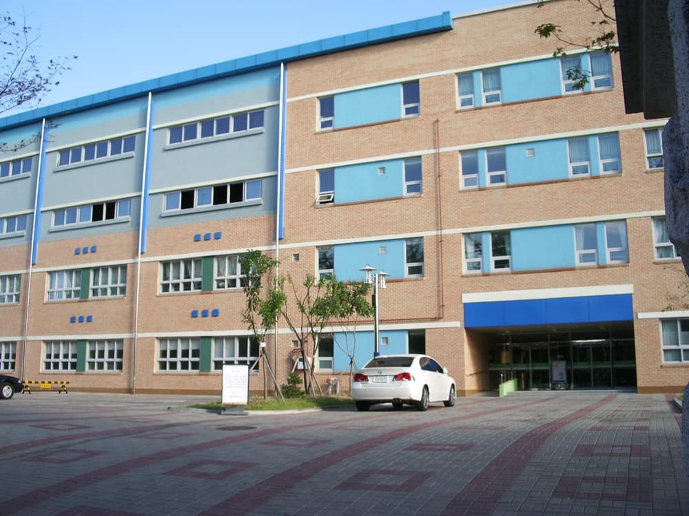 A four-storey public elementary school building in Seoul with a brick and pale blue facade, a covered blue entrance on the right and a white car parked in the paved schoolyard.