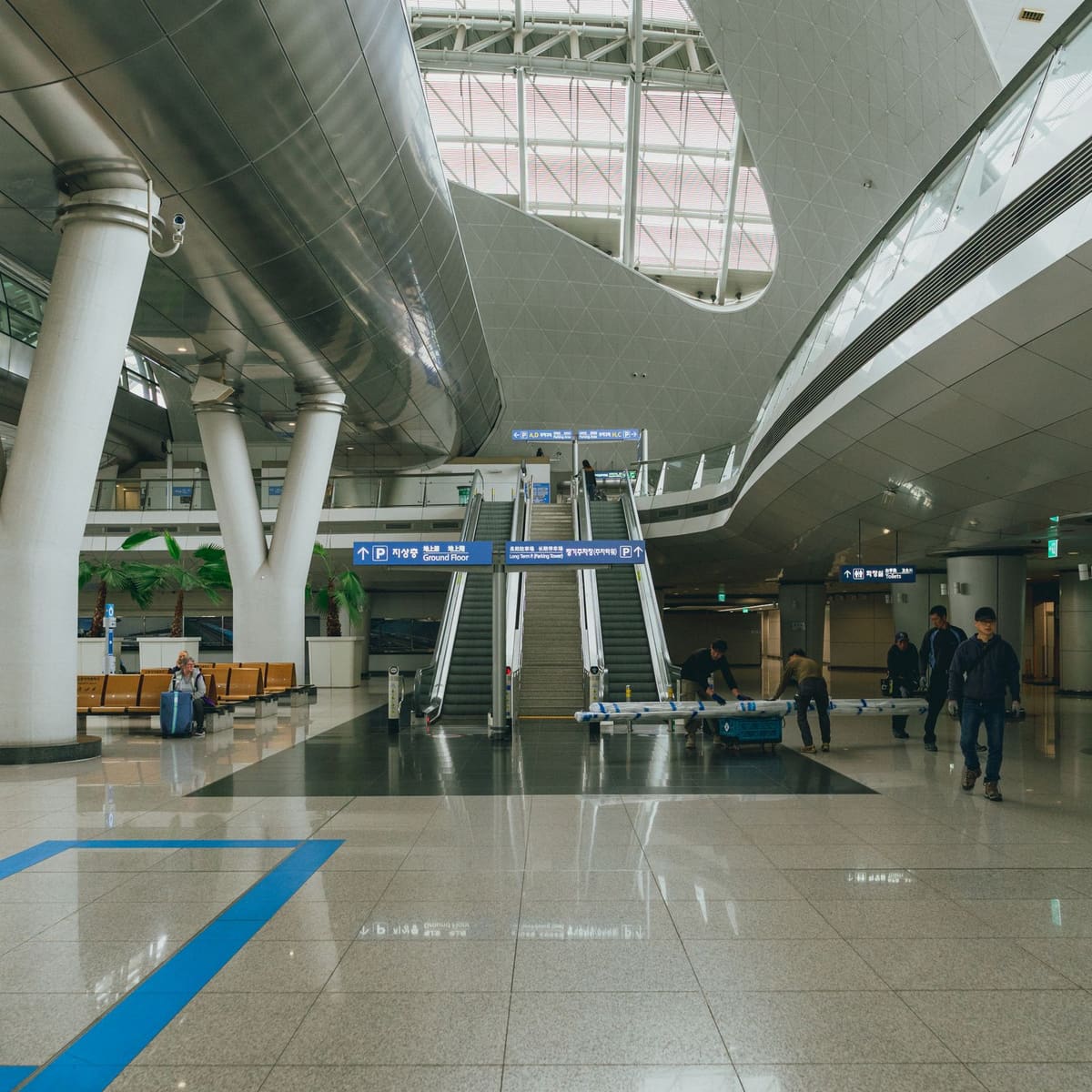 Inside the transport centre at Incheon International Airport: a high skylit atrium with curved steel ceilings, escalators under a sign for the ground floor and the long-term parking tower, a traveller seated with a blue suitcase and workers carrying a pipe across the polished floor.