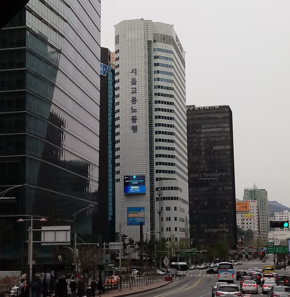 A white office tower on a Seoul avenue with the vertical Korean sign 서울고용노동청 (Seoul Regional Employment and Labor Office) and a large video screen on its facade, between glass towers, with buses and cars at a crosswalk in front, on an overcast day.