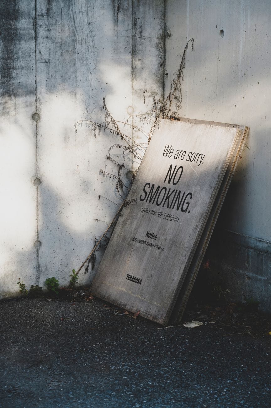 A weathered wooden board leaning against a concrete wall reads in English "We are sorry. NO SMOKING." and in Korean that smoking is banned both indoors and outdoors and that smoking brings a ₩100,000 fine; it carries the name of the Korean coffee roaster Terarosa.
