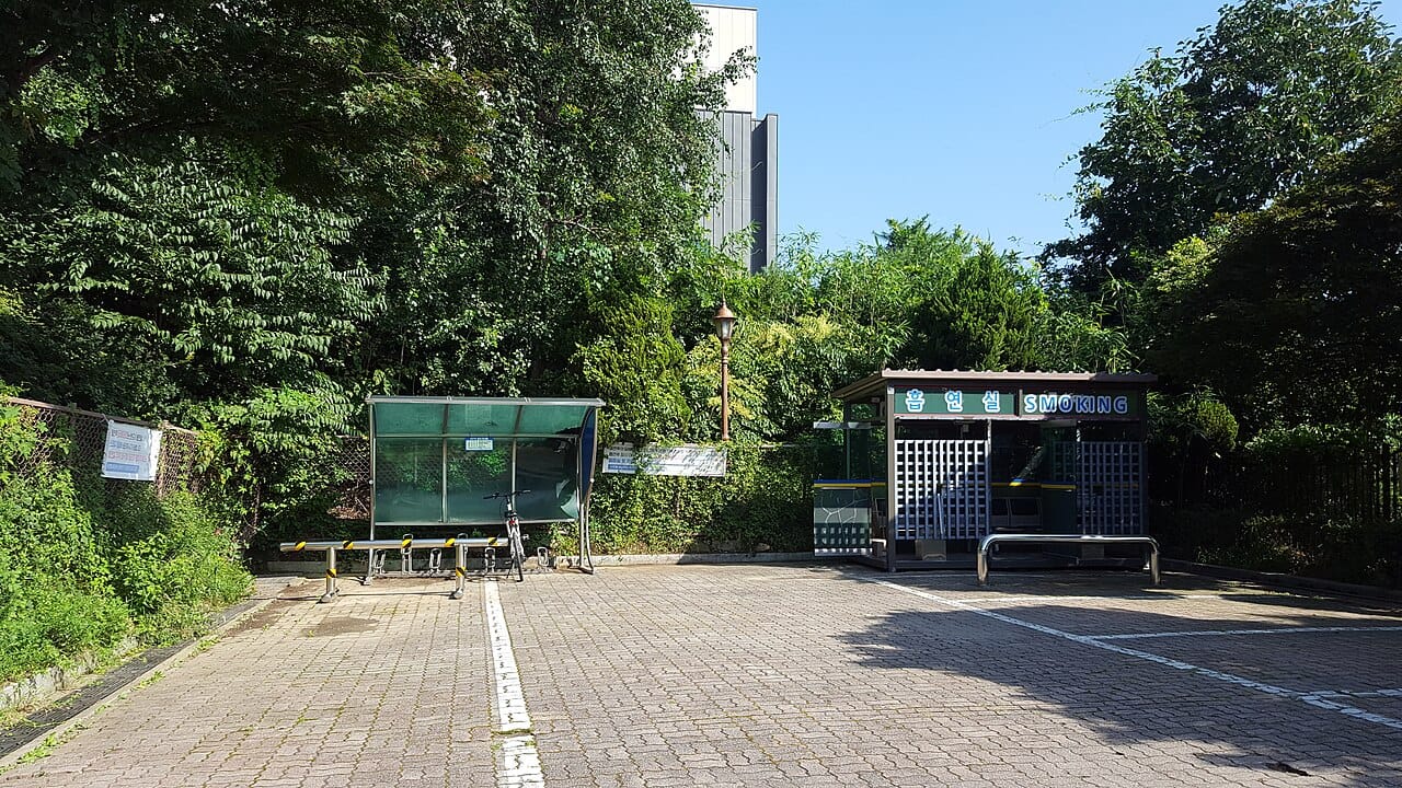 A paved car park beside Anyang Manan Library in Gyeonggi Province, with a bicycle shelter on the left and, on the right, a small roofed outdoor booth with lattice walls and a sign reading 흡연실 SMOKING.