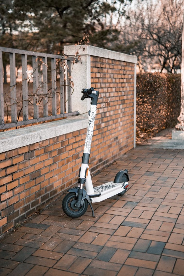 A white electric kick scooter with black graphics, parked upright on its kickstand on a red-brick pavement beside a low brick wall and railing, with bare winter trees behind.