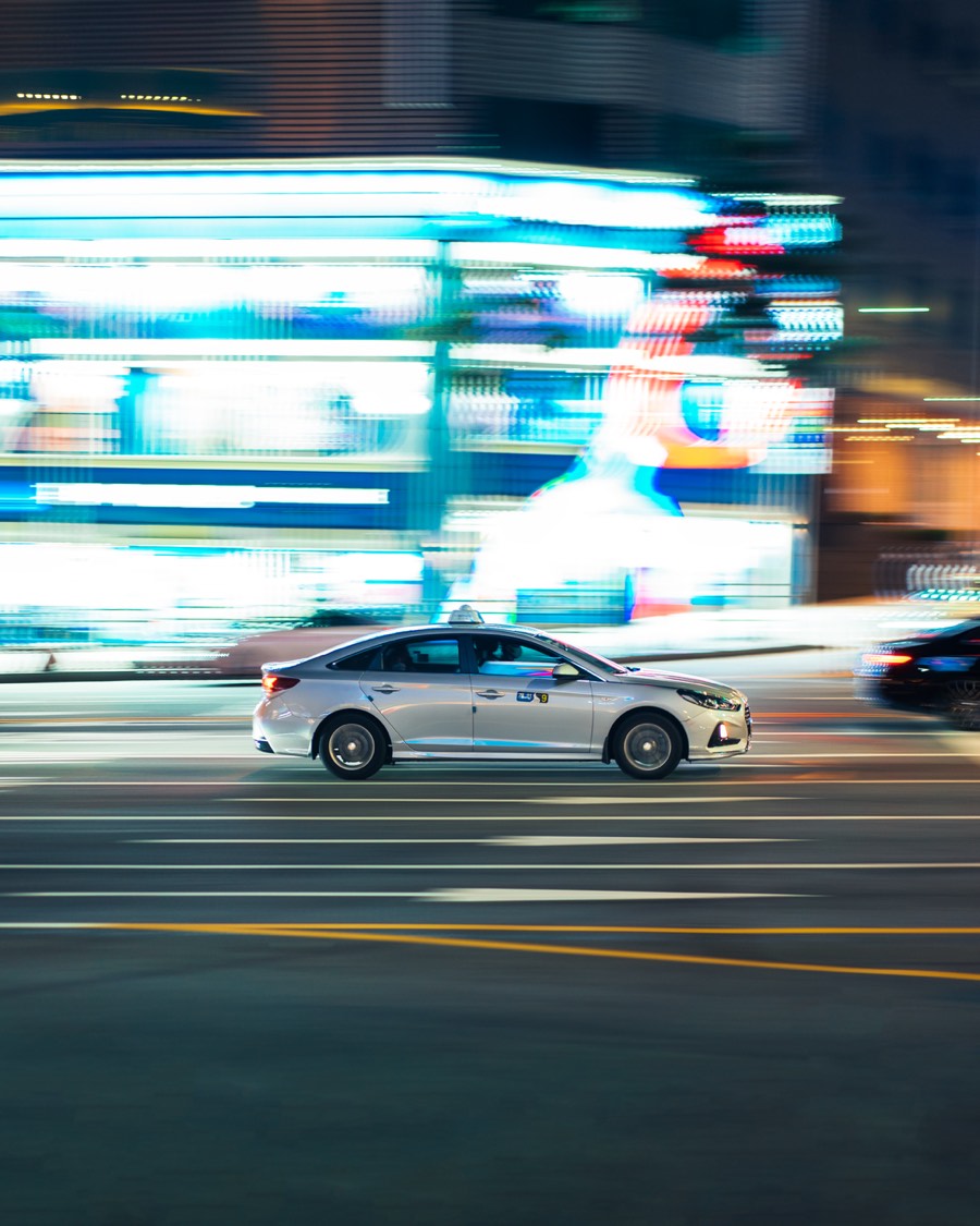A white Hyundai Sonata taxi with a roof sign, panned in sharp focus against a blur of neon shop lights on a Seoul street at night, with lane markings and a yellow centre line in the foreground.