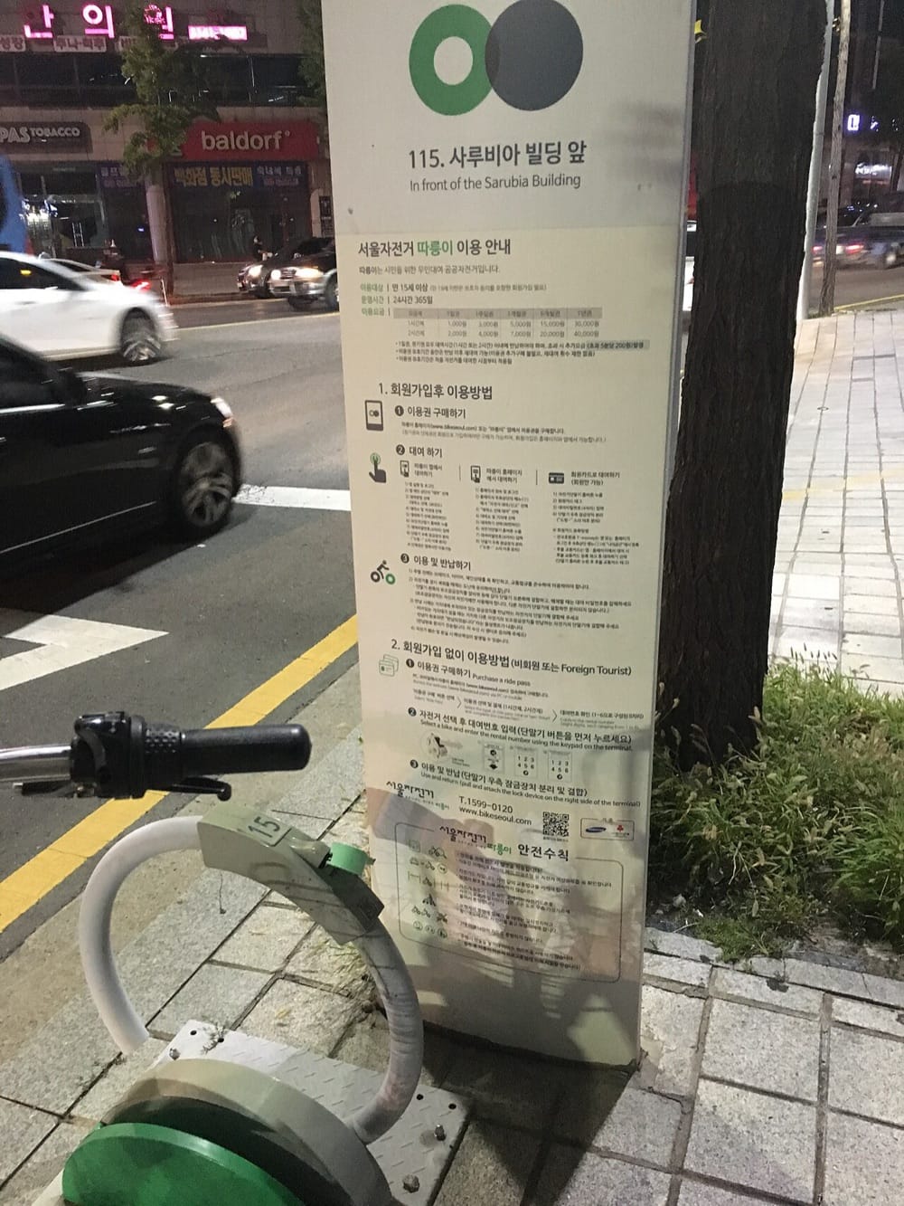 A Seoul Bike (Ttareungi) station pillar at night on a city street, its numbered sign reading "115. In front of the Sarubia Building" above a Korean and English user guide, with a docked bike's handlebar and lock cable in the foreground and cars passing on the road behind.