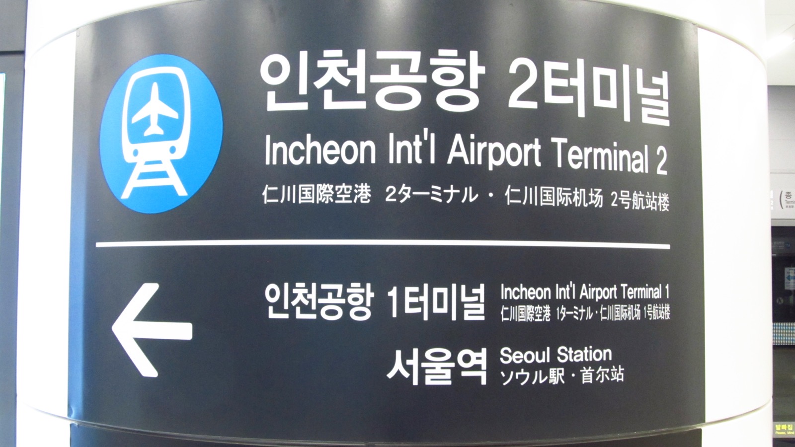 A dark station sign on a pillar at Incheon International Airport Terminal 2 Station, with a blue airport-railroad symbol, the station name in Korean, English, Japanese and Chinese, and an arrow pointing to trains for Incheon Airport Terminal 1 and Seoul Station.