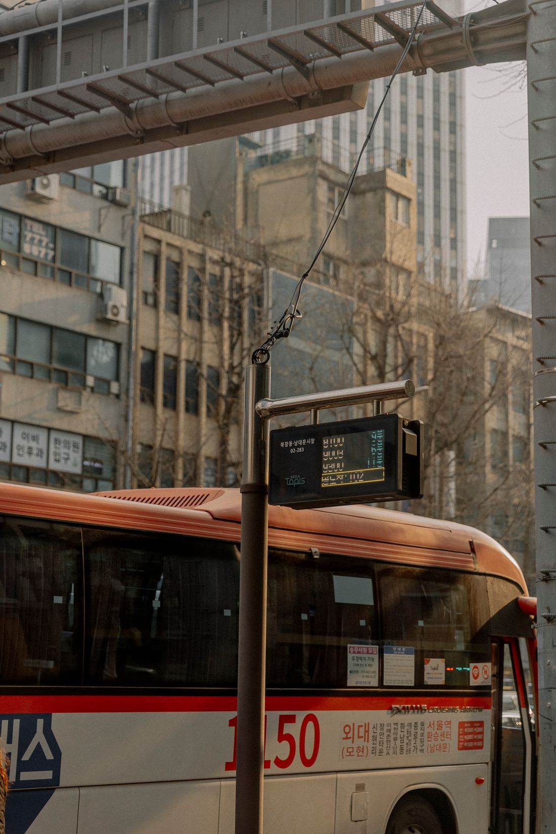 A Seoul street with a red wide-area express bus on route 1150 pulling in beside a bus-stop pole whose digital panel shows arriving buses, with older office buildings and a tower behind.