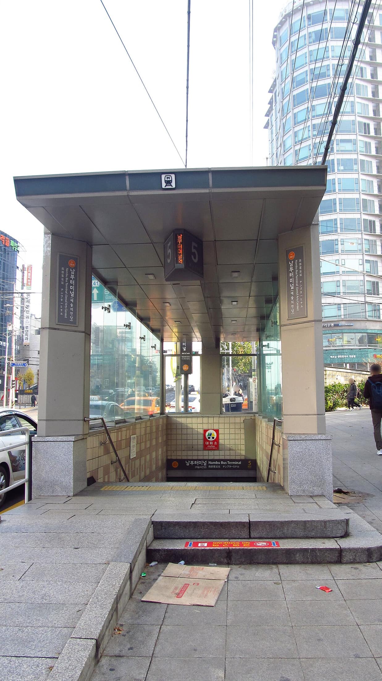 The number 5 entrance to Nambu Bus Terminal Station on Seoul Metro Line 3: a covered stairway with glass sides, an orange Line 3 badge and signs reading 남부터미널 (예술의전당) Nambu Bus Terminal (Seoul Arts Center), with a curved glass office tower behind it on a clear autumn day.