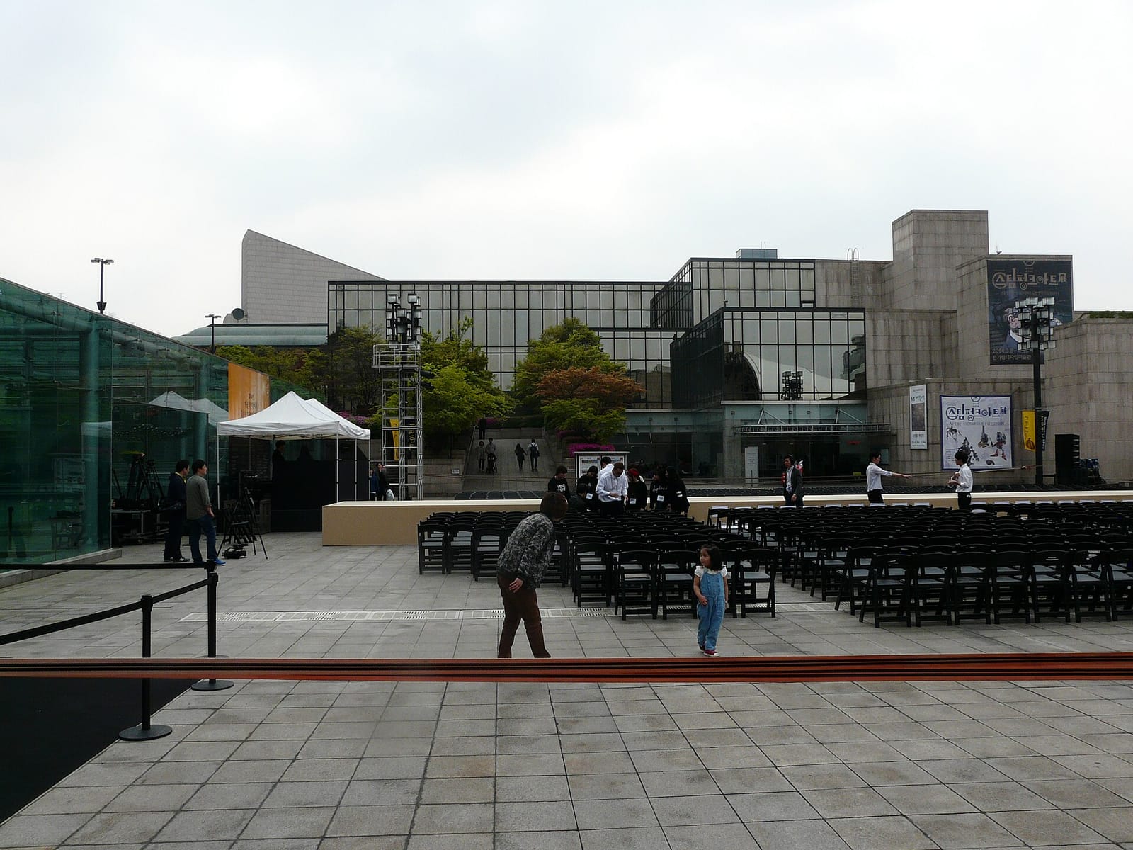 The plaza inside the Seoul Arts Center on an overcast spring day: grey paving, rows of black folding chairs being set up in front of a low stage, a white tent, and glass-and-stone buildings behind with exhibition banners and trees.