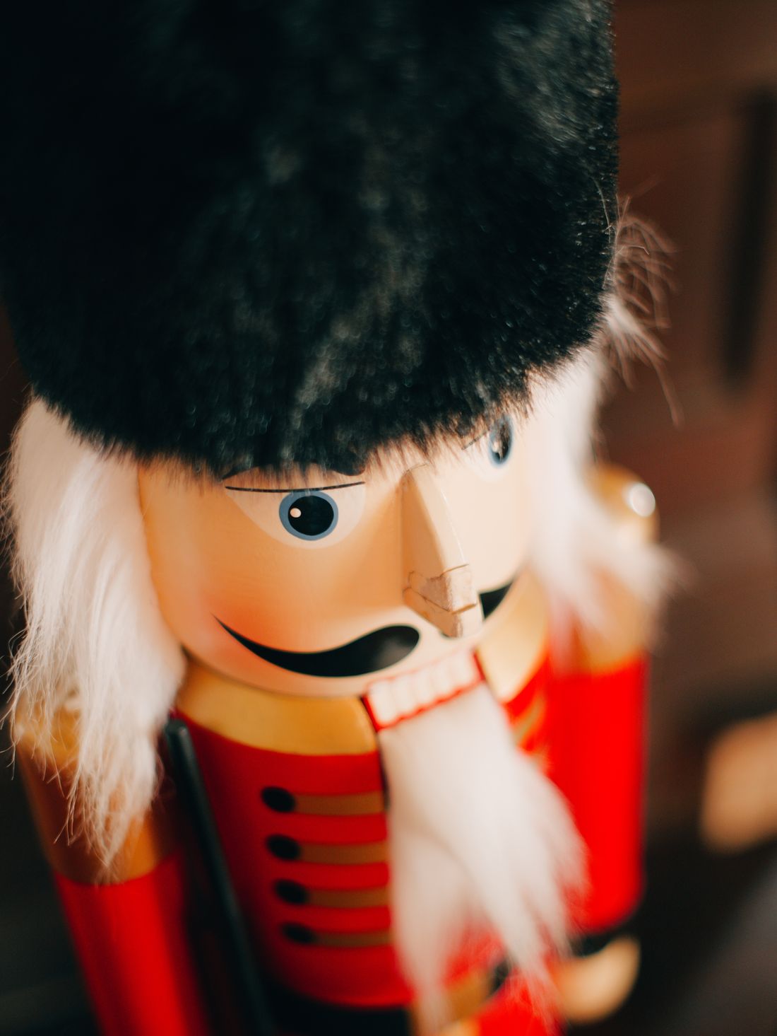 Close-up of a traditional wooden nutcracker figure with a tall black fur hat, painted blue eyes and black moustache, a white fur beard and a red jacket with gold trim, against a soft dark background.