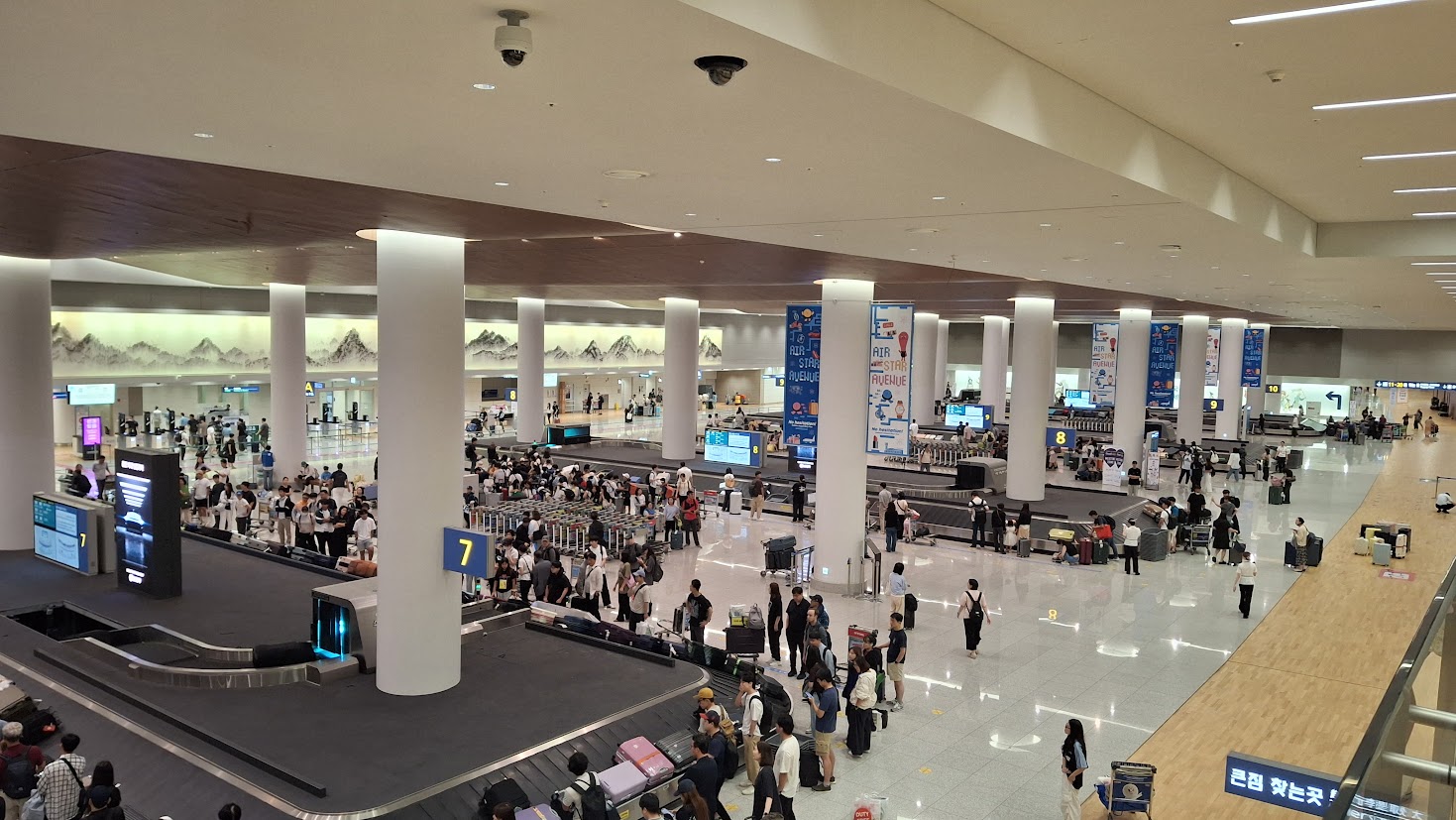 The Incheon Airport Terminal 2 baggage hall seen from above: rows of white pillars, baggage carousels marked 7 and 8 with crowds of travellers and luggage trolleys, a mountain-landscape mural along the far wall and a blue sign in Korean pointing to oversized baggage.
