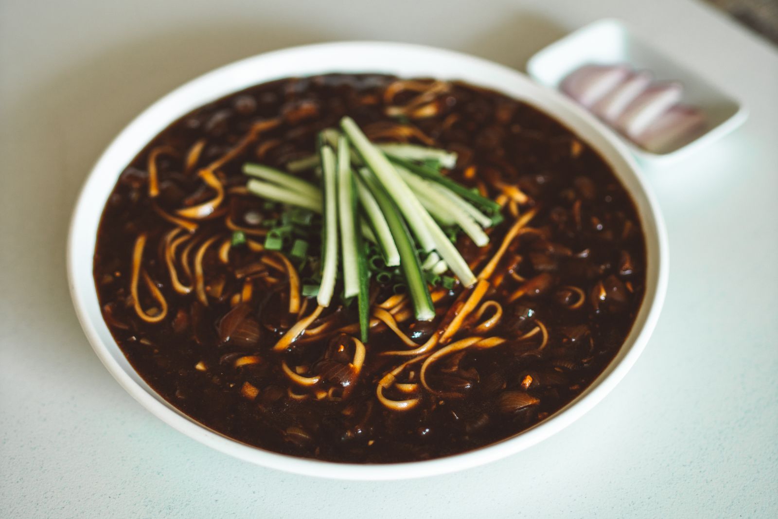 A bowl of jajangmyeon, wheat noodles in a thick glossy black-bean sauce topped with thin cucumber strips and spring onion, with a small dish of sliced raw onion behind it.